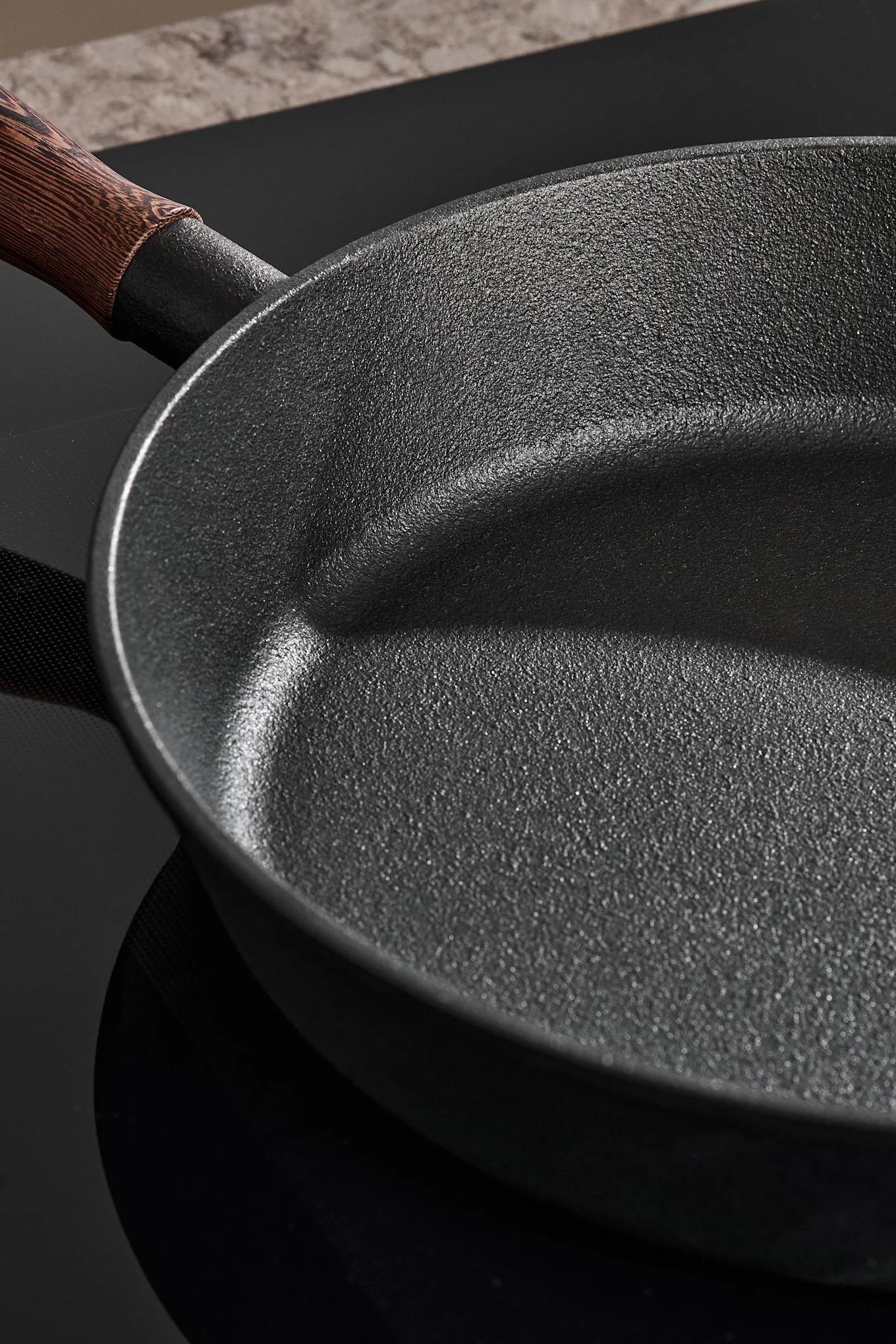 Close-up of a dark, textured frying pan with a wooden handle on a black stovetop.